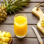 Pineapple juice in glass closeup near sliced fruit on wooden background.